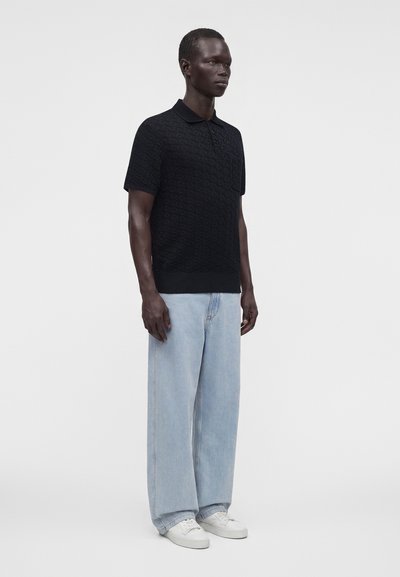 Homme debout portant un polo noir texturé à manches courtes, un jean large bleu clair et des baskets blanches sur un fond uni.