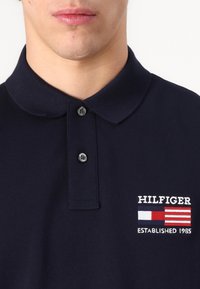 Marine polo skjorte laget av bomull, med en to-knapps placket, krage, brodert "HILFIGER" logo, og flaggdesign med røde, hvite og blå detaljer.