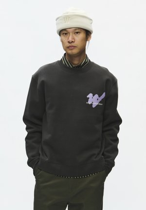 Mann trägt weiße Mütze und dunkles Sweatshirt mit lila Grafik und Text, steht mit den Händen in den Taschen vor schlichtem Hintergrund.