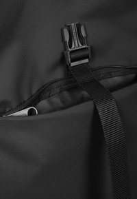 Schwarze Stofftasche mit einem Reißverschlussöffnungs, ausgestattet mit einer schwarzen Kunststoffschnalle und einem gewebten Tragegurt, der eine strukturierte Oberfläche betont.