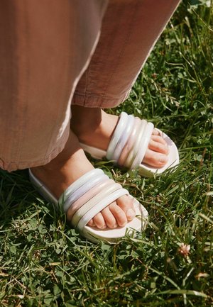 Pėdos dėvinčios baltas basutes su storomis pastelinėmis juostelėmis, stovinčios ant žolės, derinamos su šviesiai rožiniais kelnėmis.