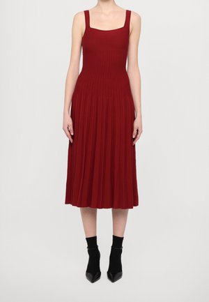 Donna che indossa un vestito midi senza maniche, a coste e plissettato di colore rosso scuro con scollo quadrato, abbinato a stivaletti neri e calzini, in piedi su sfondo neutro.