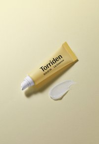 Gul tube med Torriden Solid In Lip Essence med en hvid applikator, ved siden af en klar, glansfuld stribe af lip essence på en lys baggrund.