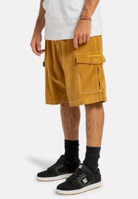 Buks i kordfløjl i sennepsgul med cargo-lommer. Parret med sorte sneakers og ribbede sokker. Enkel hvid skjorte.