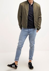 Chaqueta bomber verde oliva con cuello acanalado, camiseta polo azul marino, jeans azul claro y zapatillas negras con rayas blancas.