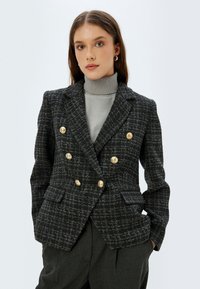 Zwart en grijs geblokte blazer met een dubbel borst ontwerp, gouden knopen en twee voorzakken, van textuurstof, gepresenteerd op een model.