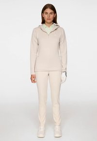 Beige zip-up tröja med huva, kombinerad med matchande slimfitbyxor. Outfiten innehåller en ljusgrön skjorta med krage under. Vit golfhandske.