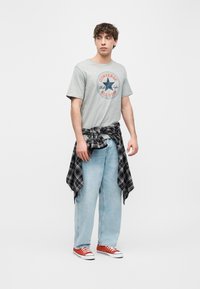 Νέος άντρας που φοράει γκρι μπλουζάκι Converse All Star, ανοιχτόχρωμο μπλε τζιν, κόκκινα αθλητικά παπούτσια και πουκάμισο καρό δεμένο γύρω από τη μέση, στέκεται μπροστά σε λευκό φόντο.