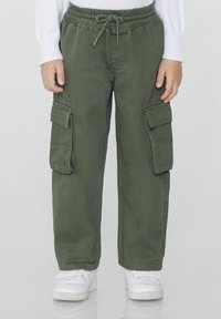 Dítě oblečené v olivově zelených cargo kalhotách s páskem na stažení a bílých teniskách, stojící na jednobarevném pozadí.