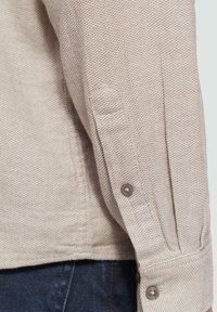 Karamelkleurig, tekstuur-rijk lang mouwshirt met een geweven patroon, voorzien van een knoopmanchet en subtiele stikdetails op de mouwnaad.
