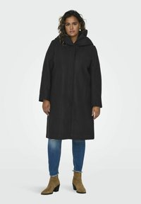 Manteau noir à capuche avec un design droit et long jusqu'aux genoux. Il présente des manches longues et une texture lisse, associé à un jean bleu clair et des bottines à talons beige.