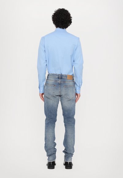 Lichtblauwe lange mouw blouse met een gladde textuur, gecombineerd met vervaagde blauwe jeans en zwarte schoenen, gezien vanaf de achterkant.