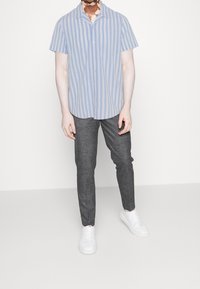 Osoba ubrana w niebiesko-białą pionowo paskowaną koszulę z krótkim rękawem, szare w kratkę spodnie i białe sneakersy, stojąca na białym tle.