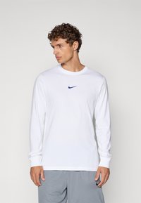 Hvid langærmet Nike t-shirt med marineblå logo, rund hals, ribbede manchetter og glat stof. Sammen med grå shorts.