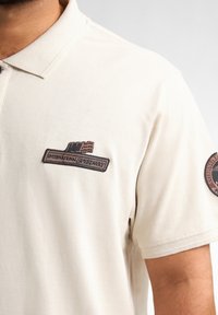 Beige polo shirt gemaakt van een textuurstof, met een patch met "INTERNATIONAL SPEEDWAY" en een vlagontwerp op de borst en een ronde mouwpatch.