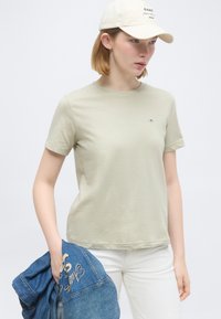 Νεαρή γυναίκα με μπεζ μπλουζάκι και καπέλο, κρατώντας ένα μπλε τζιν μπουφάν, στέκεται μπροστά σε απλό φωτεινό φόντο.