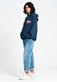 Felpa blu navy con "Amour" ricamato in rosso, dotata di una tasca frontale e cappuccio con cordino. Abbinata a jeans azzurri arrotolati e sneakers bianche.