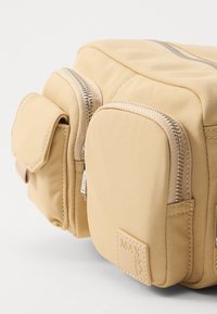 Borsa in tessuto morbido beige con molteplici compartimenti con zip, dotata di una superficie testurizzata e di una patch quadrata con logo. Include una tasca frontale.