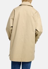 Beżowy trencz z szerokim kołnierzem, długimi rękawami i prostym krojem. Tkanina ma gładką fakturę i minimalne detale. Noszony na niebieskie dżinsy.