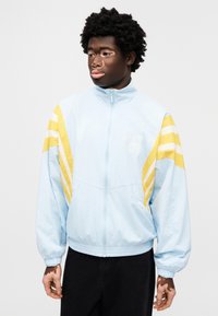 Chaqueta azul claro con rayas amarillas en los hombros, cierre con cremallera y dobladillo acanalado. El material parece liso y ligero.