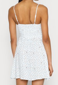 Vit klänning med blå blommönster, smockad livdel och tunna justerbara axelband. Flödande kjol som går strax ovanför knät.