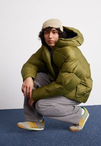 Green puffer jacket with a hood, featuring a glossy texture. The model wears gray corduroy pants and gray sneakers with yellow accents.