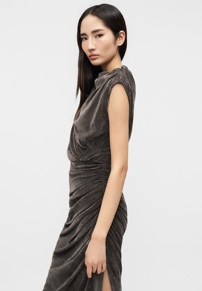 Νέα γυναίκα με μακριά σκούρα μαλλιά, που φοράει ένα αμάνικο, ρυτιδιασμένο, σκουρόχρωμο βελούδινο φόρεμα, στέκεται μπροστά σε ένα απλό φωτεινό φόντο.