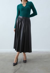 Femme portant un haut à manches longues couleur sarcelle avec un décolleté en forme de trou de serrure, une jupe midi plissée noire et des escarpins noirs à bouts pointus, debout sur un sol en béton.