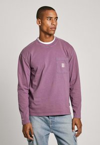 Long-sleeve purple T-shirt in soft cotton with a front pocket, rounded neckline, and subtle stitching details on sleeves.