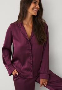 Blomme satin pyjamasæt med lange ærmer, krave og knaplukning foran. Har kontrasterende sort piping langs kanterne og manchetterne. Glat tekstur.