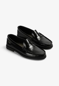 Par de mocasines de piel negra con tacones bajos y detalles cosidos, sobre un fondo blanco.