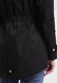 Black jacket with a cinched waist, smooth fabric, and gold-colored snap buttons. Features a slightly curved hem and clean seams.