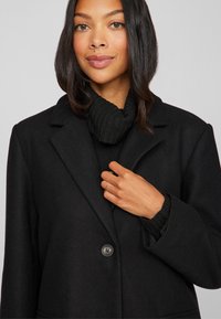 Mustlane villasegu jope, millel on sooniline kaelus, üksainus nööp kinni ja tekstuuriga must sviiter all. Lihtne, kohandatud disain.