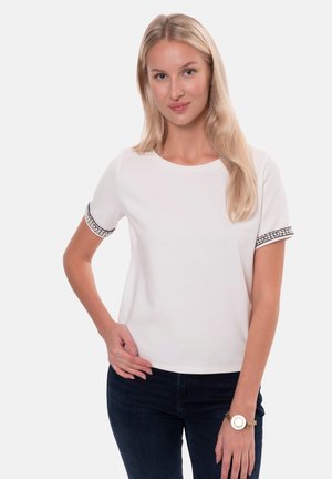 Biała koszulka z krótkim rękawem z dekoracyjnym wykończeniem rękawów, okrągłym dekoltem, noszona z ciemnymi dżinsami i bransoletką w złotym odcieniu. Gładka faktura materiału.