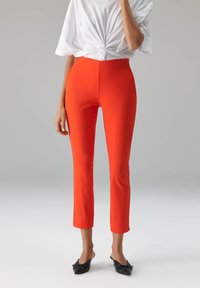 Rode nauwsluitende broek met een gladde textuur, nette lengte en een hoog getailleerd ontwerp. Draagt een wit geknoopt topje en zwarte platte schoenen.