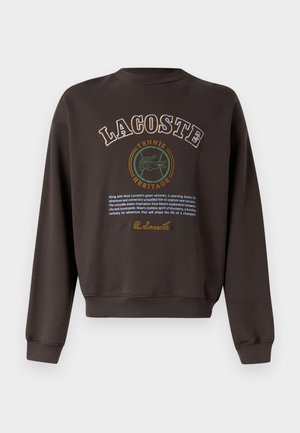 Temno rjava Lacoste majica z napisom "LACOSTE TENNIS HERITAGE", zelenim logotipom krokodila in odstavkom o René Lacostovi inspiraciji.