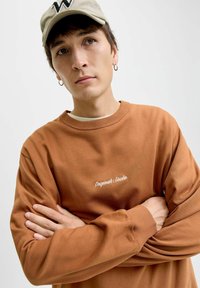 Felpa marrone con polsini e scollatura a coste, con sopra ricamato in bianco il testo "Originals Studio." Le braccia del modello sono incrociate.