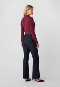 Kobieta stojąca bokiem, ubrana w dopasowaną bordową bluzkę z długim rękawem, ciemnoniebieskie jeansy typu bootcut i bordowe buty na niskim obcasie na szarym tle.