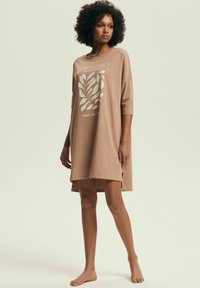 Kobieta z kręconymi włosami, ubrana w luźną, beżową sukienkę do kolan w botaniczny wzór, stojąca boso na jednolitym jasnym tle.