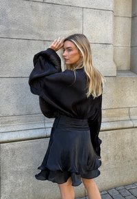 Melnas satīna blūze ar pūkainām piedurknēm apvienota ar slāņotu melnu svārku. Gluda tekstūra un plūstošs dizains ar viļņveida apakšmalu detaļām.