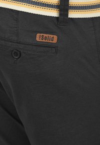 Shorts en tissu noir avec une ceinture tissée arborant des rayures jaunes et bleues. Comprend une petite étiquette en cuir marron avec « !Solid » sur la poche arrière.