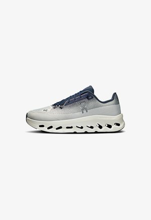 Lichte hardloopschoen in grijs en marineblauw. Kenmerkt zich door een ademend bovenwerk van mesh, een dikke witte zool met uitsparingen en platte blauwe veters.