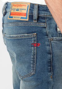 Kék denim farmernadrág bőr címkével, narancssárga varrással és piros hímzett logóval a hátsó zseben. Normál szabás, közepes mosás.