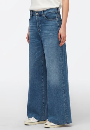 Persona con vaqueros azules de pierna ancha, cintura alta y dobladillo sin rematar, combinados con zapatillas blancas y una camiseta blanca.