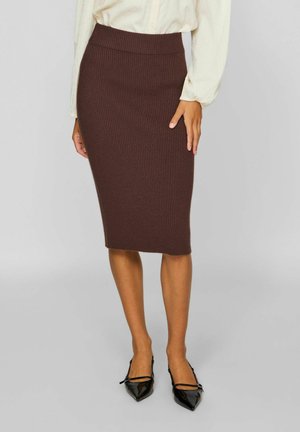 Femme portant une jupe midi en maille marron côtelée ajustée, un chemisier beige et des talons noirs pointus, debout devant un fond uni.