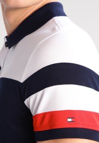 Polotröja med marinblå, vit och röd rand. Har krage, korta ärmar och en logotypdetalj på ärmen. Tillverkad av bomull med strukturerad yta.