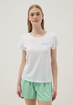 Mujer joven con camiseta blanca con texto azul "soleil d'été" y pantalones cortos a rayas verdes, de pie contra un fondo claro y liso.