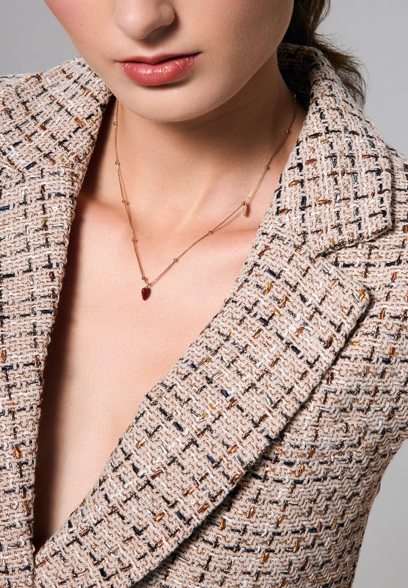 Złoty naszyjnik z czerwoną zawieszką w kształcie serca, noszony na beżowej marynarce o teksturowanej powierzchni z wielokolorowymi włóknami, z głębokim dekoltem w kształcie litery V.