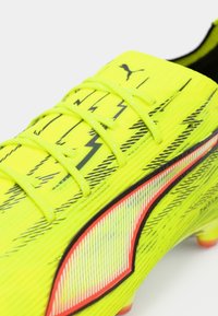 Lyse gul fodboldstøvle med tekstureret overdel, sorte lynmønstre, reflekterende detaljer og sorte snørebånd. Udstyret med knopper til fast underlag.