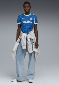 Niebieska koszulka sportowa z teksturowanym wzorem, krótkie rękawy i zielone akcenty, zestawiona z jasnoniebieskimi dżinsami o szerokich nogawkach i białymi sneakersami. Jasnoszara kurtka przewiązana w talii.